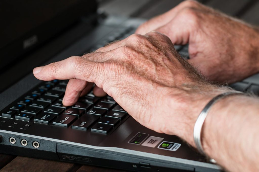 Valokuvassa vanhuksen kädet näpyttelee kannettavaa tietokonetta.