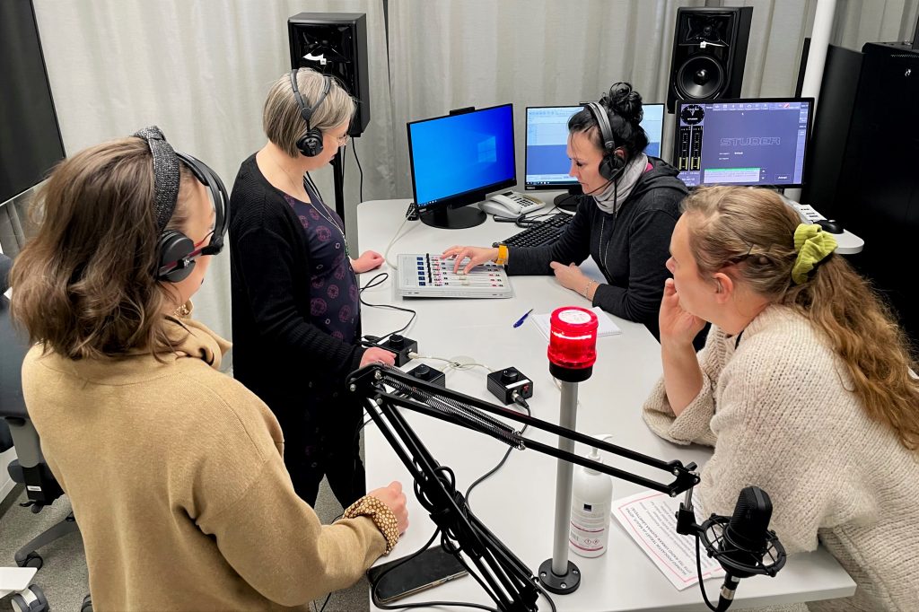 Valokuva radiostudiosta, jossa neljä ihmistä on pöydän ääressä ja yksi heistä säätää äänen tasoa.