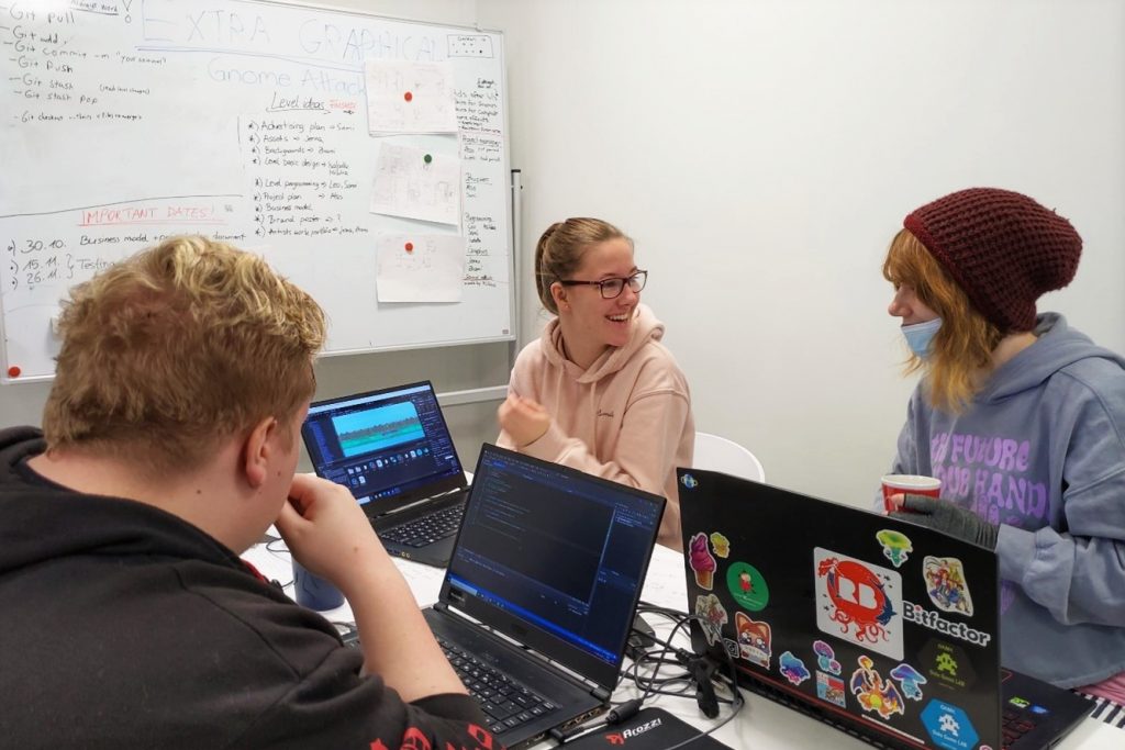 Valokuva, jossa kolme ihmistä istuu tietokoneiden ääressä ja hymyilee.
