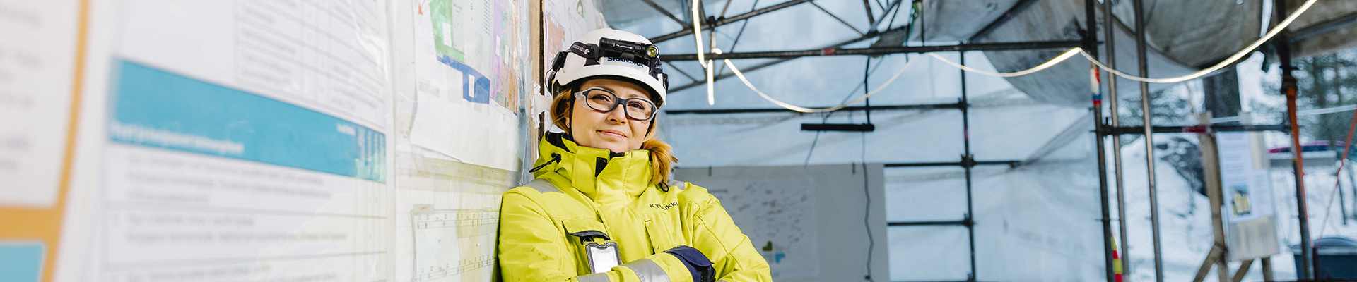 Nainen huomiovaatteissa ja suojakypärässä hymyilee työmaakatoksessa