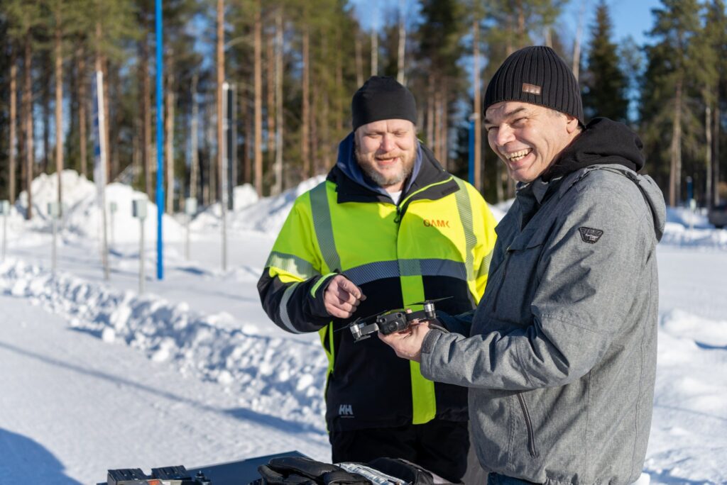 Kaksi hymyilevää miestä droonin kanssa aurinkoisessa kevättalven säässä