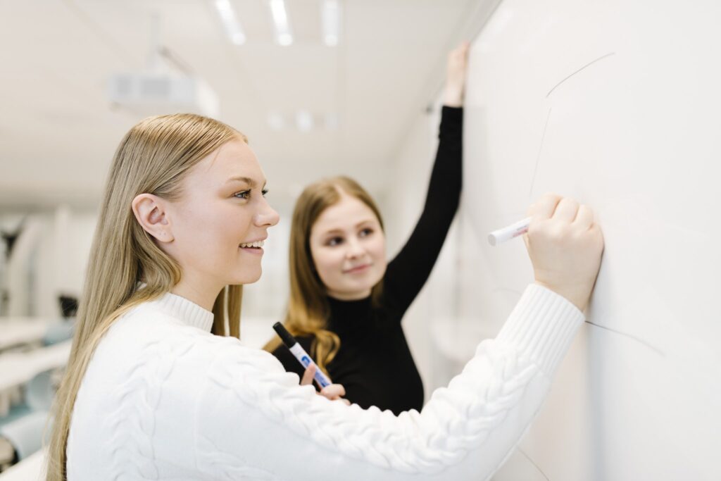 Nuori naisopiskelija piirtää valkotaululle tussilla ja toinen nuori nainen seuraa vieressä.